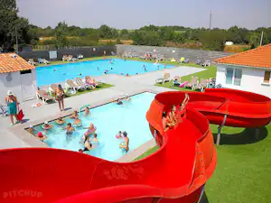 Großes Camping-Schwimmbad mit Wasserrutsche, Liegen und Rasenfläche, daneben Gebäude mit orangefarbenem Dach.