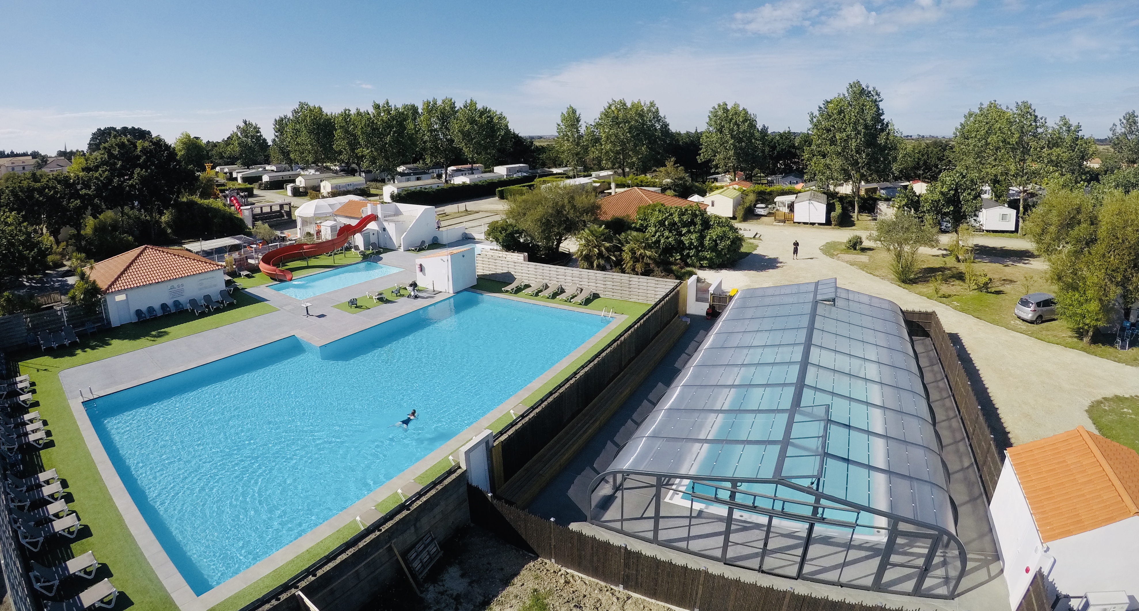 Großes Schwimmbecken mit Liegefläche, daneben ein Saunabereich und Stellplätze mit Wohnwagen, umgeben von Bäumen.