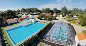 Großes Schwimmbecken mit Liegefläche, daneben ein Saunabereich und Stellplätze mit Wohnwagen, umgeben von Bäumen.