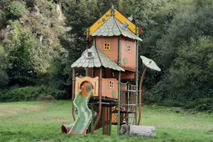 Spielhaus für Kinder mit Rutsche und Klettergerüst auf grünem Rasen vor dichtem Wald.