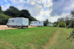 Zwei Wohnwagen auf einer grünen Campingwiese, daneben ein Zelt und Hecken, im Hintergrund Bäume und ein Weg.