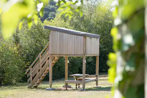 Holzspielhaus auf Stelzen im Campinggarten, mit kleiner Bank und Tisch, umgeben von Büschen und Bäumen.