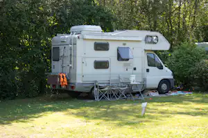 Wohnmobil mit ausgeklappter Markise und Vorzeltstühlen am Stellplatz im Grünen, umgeben von Bäumen und Rasen.