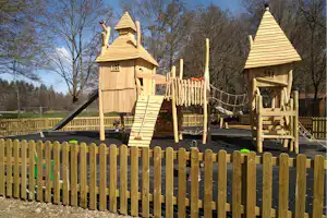 Holz-Spielhaus mit Rutsche, Holzgeländer und Spielbereich, im Hintergrund Bäume und Wiese am Campingplatz.