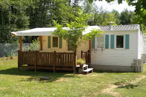 Mobilheim mit Terrasse, Markise und grünen Fensterläden, Holzsichtschutz und Garten mit Bäumen im Hintergrund.