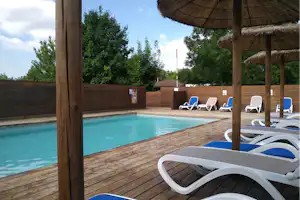 Camping-Pool mit türkisfarbenem Wasser, Holzdeck und Liegen unter einem Strohdach-Pavillon, dahinter hohe Hecken.