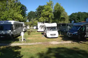 Wohnmobile auf einem Campingplatz zwischen Bäumen, mit Grasfläche, Stellplätzen und Stromanschluss am Boden.