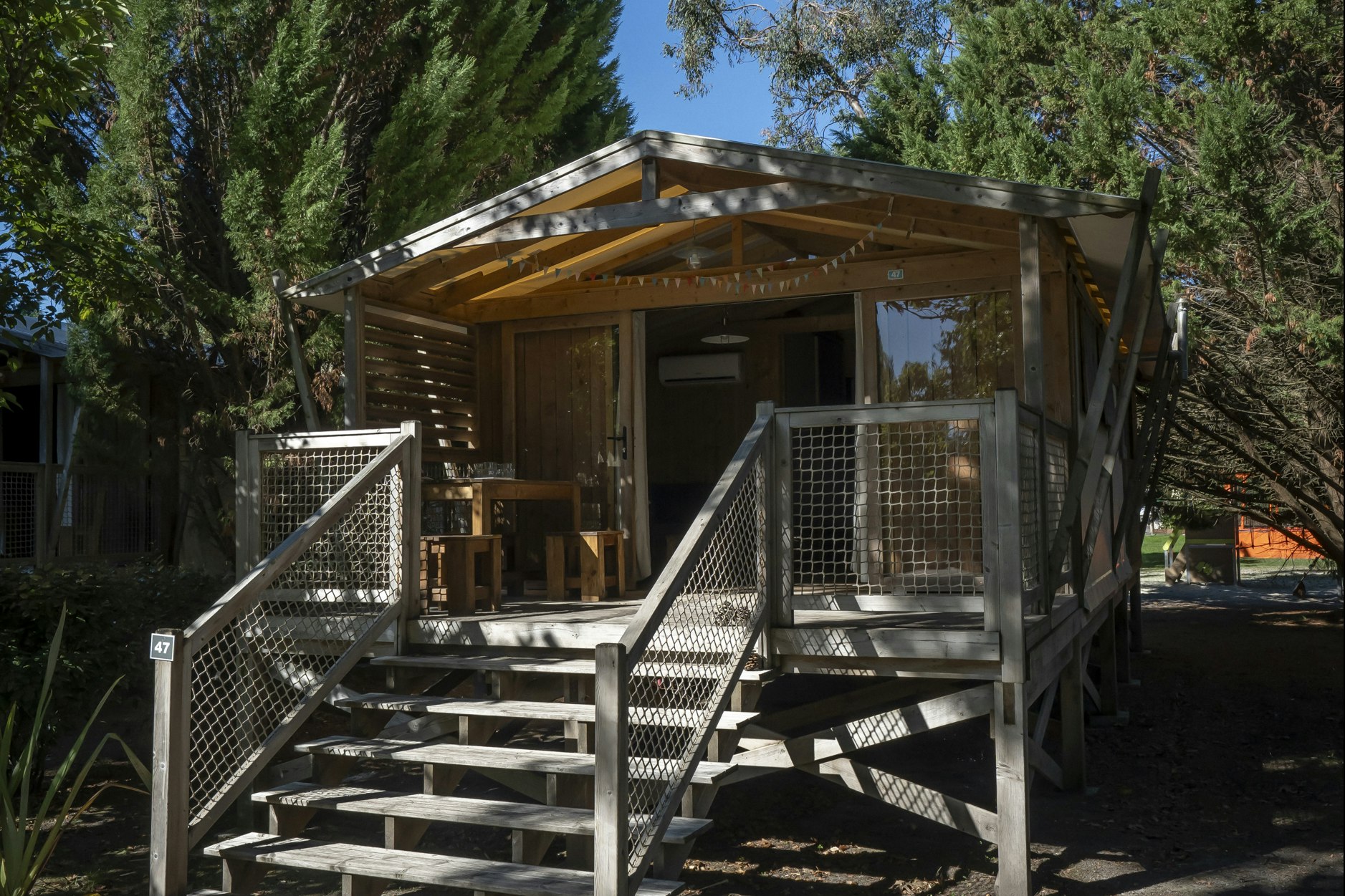 Holzunterstand mit Veranda und Treppe, umgeben von hohen Bäumen, auf einem Campingplatz mit Kiesweg und Sitzbereich.
