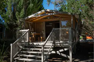 Holzunterstand mit Veranda und Treppe, umgeben von hohen Bäumen, auf einem Campingplatz mit Kiesweg und Sitzbereich.