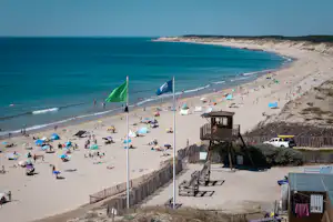 Breiter Sandstrand mit vielen Liegen und Sonnenschirmen, Blick auf das Meer und einen Zaun mit Treppe am Rand.