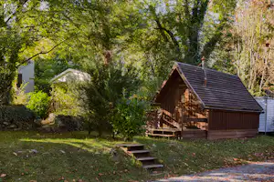 Holzhütte auf einem Campingplatz im Grünen, mit Holztreppe, Sitzbereich und umgebenden Bäumen im Hintergrund.