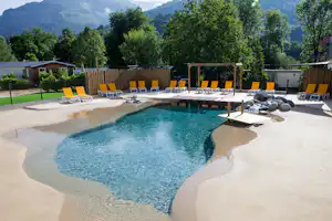 Schwimmbecken mit blauem Wasser, Holzterrasse und gelben Liegestühlen, umgeben von Bäumen und Bergkulisse.