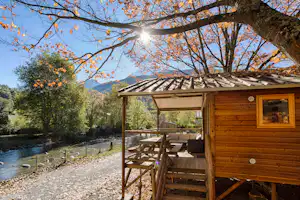 Holzhütte mit Terrasse und Sitzbank am Fluss, umgeben von Bäumen und Bergen im Hintergrund.