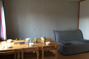 Wohnbereich mit grauem Sofa und Holztisch, Teekanne und Tassen auf dem Tisch vor einer hellen Holzvertäfelung.