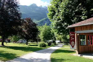 Bistro am Campingplatz mit Holzfassade, Weg und Rasenfläche, Wohnwagen im Hintergrund und Berge in der Ferne.