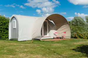 Weißes Zelt mit braunem Eingang, Holzterrasse und zwei pinken Campingstühlen im Grünen vor blauem Himmel.