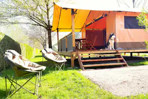 Campingzelt mit Holzterrasse, Stühlen und Tisch, daneben zwei Campingstühle im Gras vor Hecken und Bäumen.