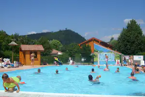 Großes Camping-Schwimmbad mit Holzgebäude, Rutsche und Liegen, im Hintergrund Bäume und ein bewaldeter Hügel.