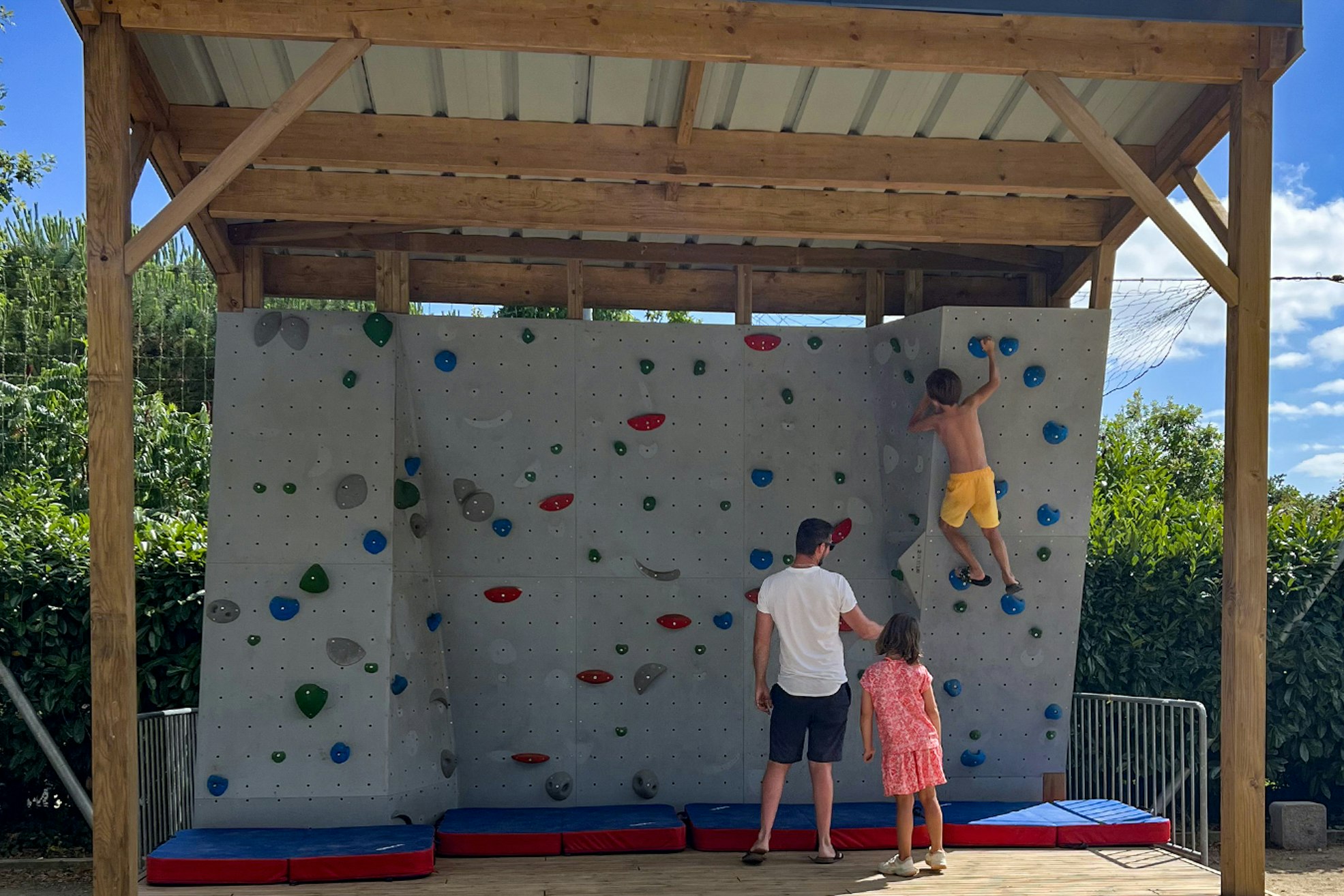Kletterwand im Campingbereich unter Holzüberdachung, mit bunten Griffen, Matten und zwei Personen am Fuß der Wand.