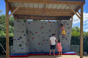 Kletterwand im Campingbereich unter Holzüberdachung, mit bunten Griffen, Matten und zwei Personen am Fuß der Wand.