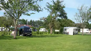 Wohnmobile und Zelte auf einer grünen Wiese, umgeben von Bäumen, mit weiteren Stellplätzen im Hintergrund.