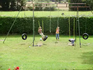 Spielplatz am Campingplatz mit zwei Schaukeln, Reifen und Rasenfläche, im Hintergrund Hecke und Zaun.
