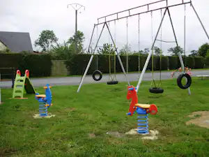 Spielplatz auf dem Campingplatz: Schaukeln mit Reifen und Federwippen, Rasenfläche, Hecken und Gartenweg im Hintergrund.