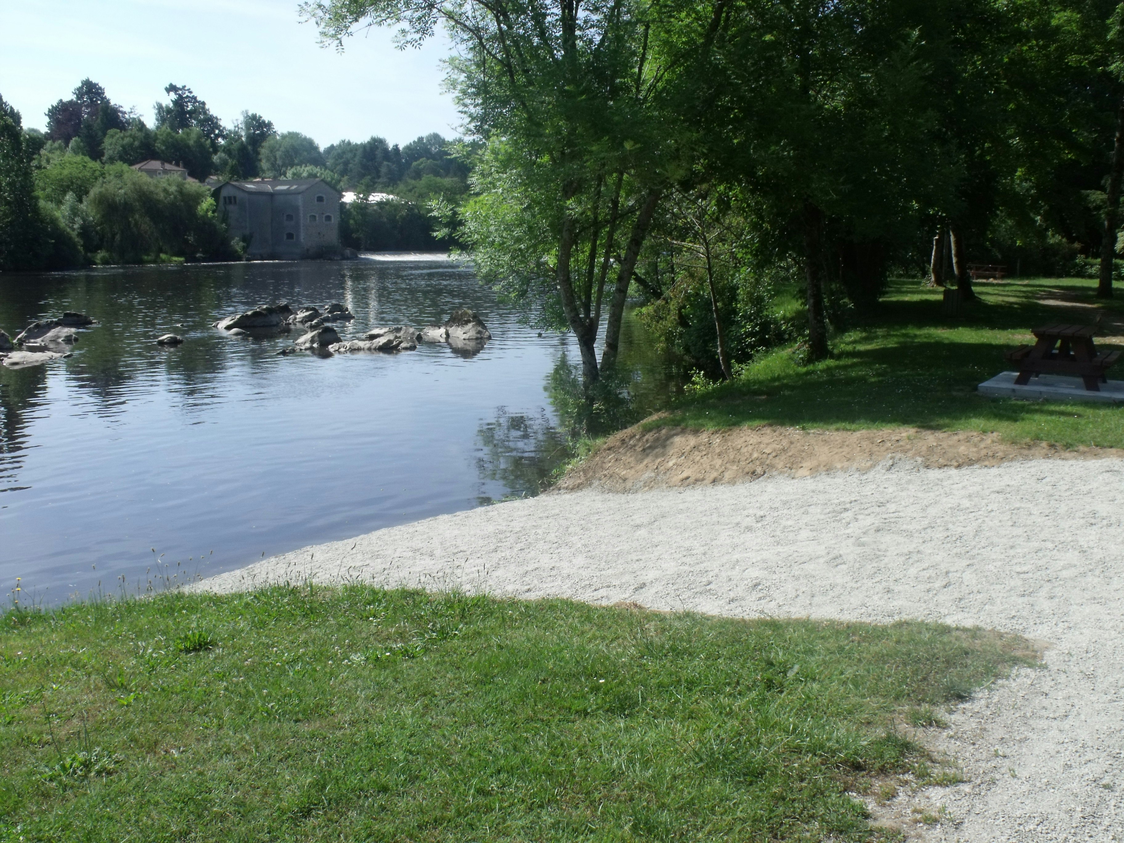 Ufer am Fluss mit Grasfläche, Baum und Bank, im Hintergrund Wasserlauf und Gebäude am Campingplatz.