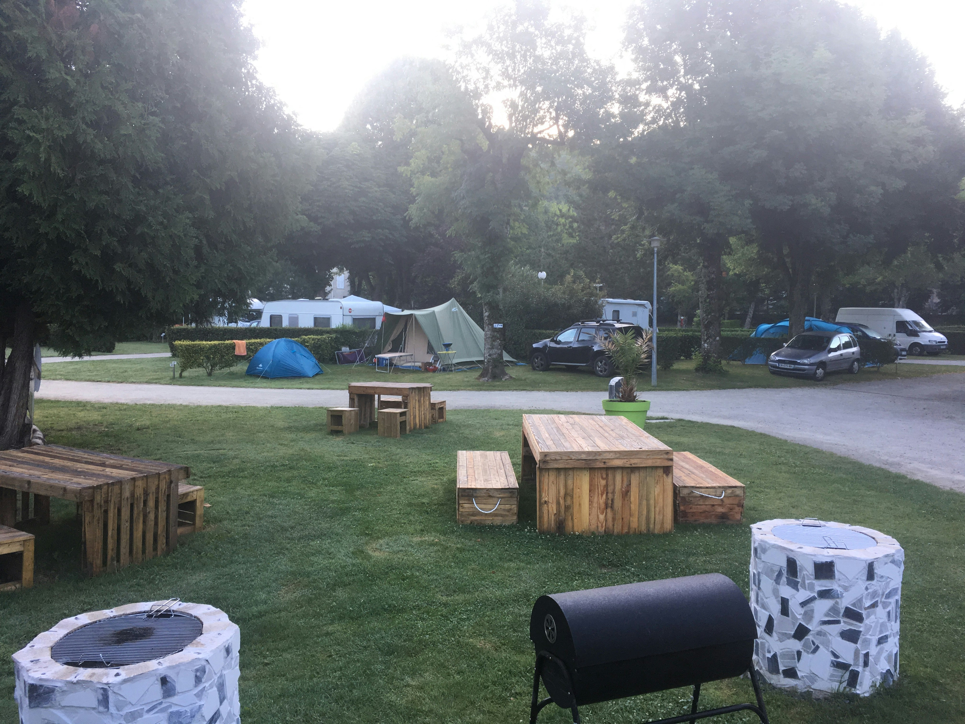 Grüner Rasen mit Sitzgelegenheiten aus Holz, Feuerstelle und Zelt sowie geparkten Autos am Campingplatz im Schatten der Bäume.