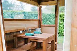 Holzterrasse am Campingplatz mit Tisch und Bank, seitlich offene Überdachung und Blick auf Hecke und Eingangstür.