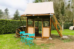 Holzunterstand am Campingplatz mit Tisch und blauen Stühlen, davor Rasen und Hecke, rechts ein Holzsteg.