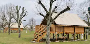 Holzhütte mit großer Veranda auf Stelzen, umgeben von Rasen und Bäumen, im Hintergrund weitere Gebäude und Wege.