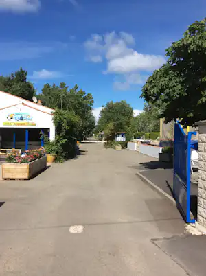 Campingplatz-Gang zwischen Gebäuden, mit blauen Toren, Pflanzkübeln und Bäumen am Rand, unter klarem Himmel.