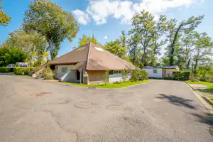 Campingbungalow mit Satteldach und Terrasse, daneben ein Wohnwagen, umgeben von Bäumen und gepflegtem Grün am Weg.