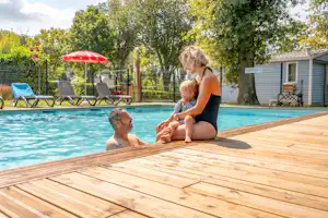 Frau und zwei Kinder am Pool auf einer Holzterrasse, im Hintergrund Liegen, Bäume und ein kleines Gebäude.