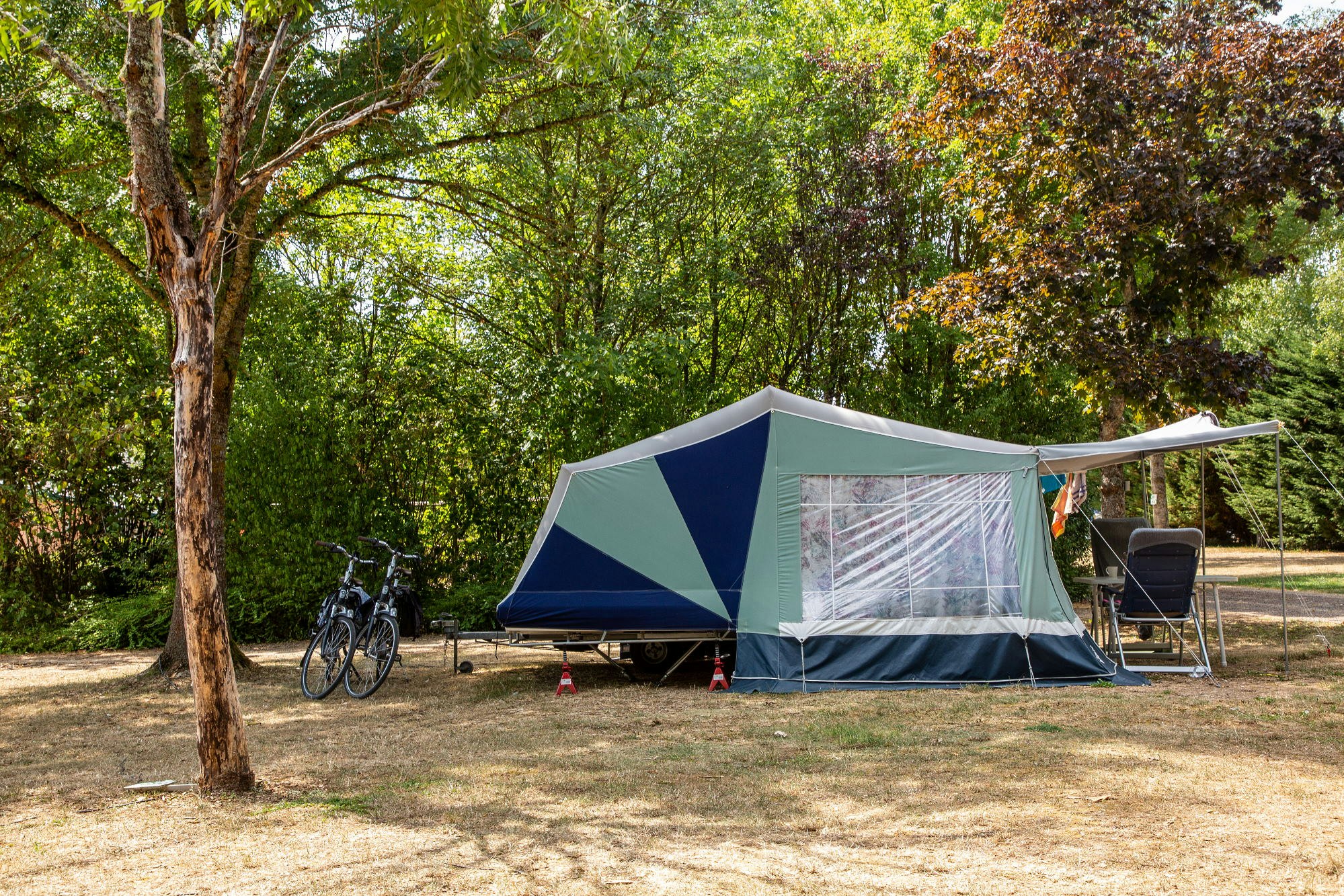 Campingzelt mit Vorzelt auf einem Stellplatz im Grünen, daneben Fahrräder und ein Stuhl am Rand der Bäume.