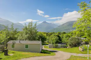 Bungalow auf einem Campingplatz im Grünen, mit Kiesweg, Hecken und Blick auf grüne Berge im Hintergrund.