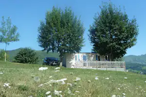 Campingwiese mit Wohnwagen, Holzsichtschutzzaun und Bäumen, im Hintergrund ein Gebäude und grüne Hügel.