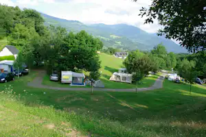 Campingwiese mit Wohnwagen und Zelten, Bäume und ein Weg, im Hintergrund grüne Berge und ein Tal.