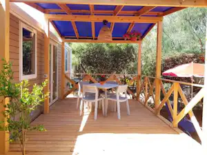 Holzterrasse am Campingplatz mit Esstisch und Stühlen unter überdachtem Pergola-Dach, daneben roter Sonnenschirm und Blick ins Grün.