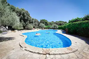 Blaues Camping-Poolbecken mit Steinrand, umgeben von Bäumen und Hecken, im Hintergrund ein klarer Himmel.