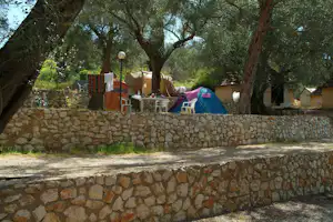 Zelte auf einem Campingplatz unter großen Bäumen, mit Steinmauer, weißen Stühlen und Tisch im Schatten.