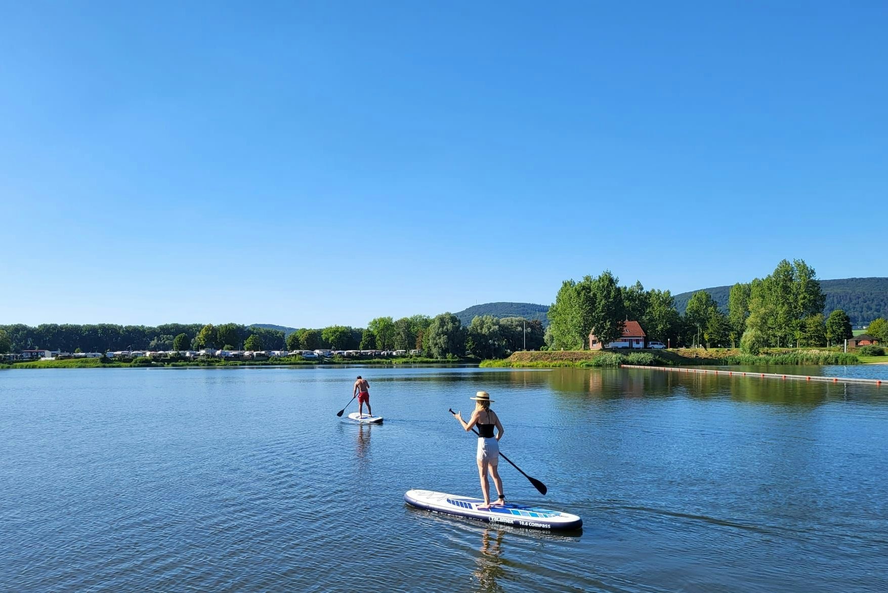 Stand-up-Paddler auf einem See, im Hintergrund Ufer mit Bäumen und Hügeln, ruhiges Wasser und blauer Himmel.