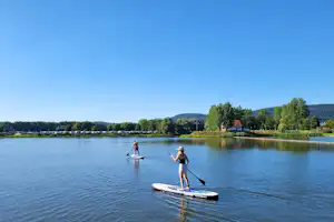 Stand-up-Paddler auf einem See, im Hintergrund Ufer mit Bäumen und Hügeln, ruhiges Wasser und blauer Himmel.