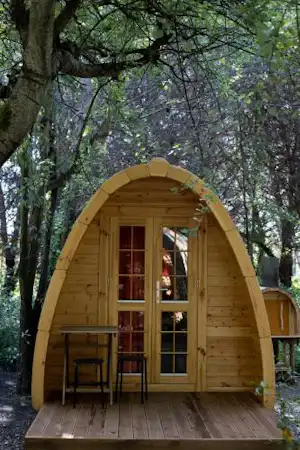 Holzhütte mit Glastür und Holzveranda im Wald, umgeben von Bäumen und einem kleinen Tisch.