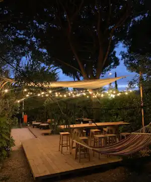Holzterrasse mit Sitzgarnituren und Hängematte, Lichterketten am Campingplatz, Bäume im Hintergrund bei Abendstimmung.