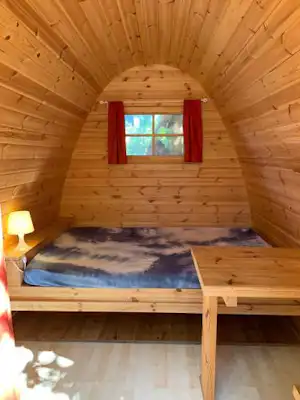 Holzsauna-Interieur mit Doppelbett, Fenster und roten Vorhängen, Holzverkleidung und Nachttischlampe links.