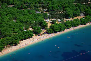 Luftaufnahme vom Campingplatz am Meer mit Strand, vielen Sonnenschirmen, Wohnmobilen und Zelten im grünen Wald.