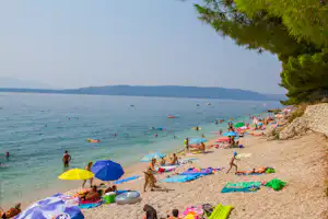 Badegäste am Kiesstrand am See, viele Schwimmtiere und Sonnenschirme, im Hintergrund Boote und bewaldete Uferhügel.