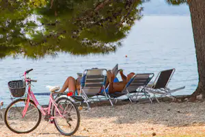Campingplatz am See: rosa Fahrrad und Liegestühle unter Bäumen am Kiesstrand, Blick aufs Wasser.